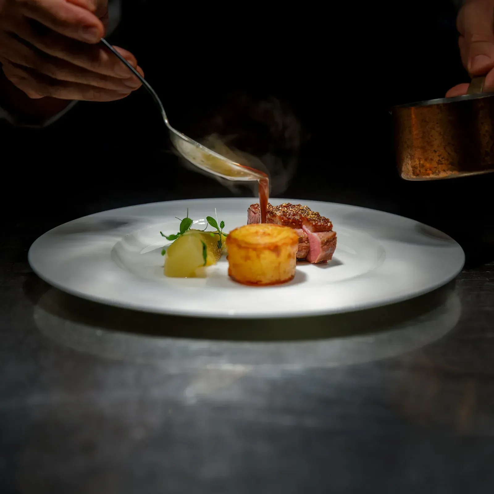 A chef pouring sauce over a plated steak with potatoes and seasonal vegetables.