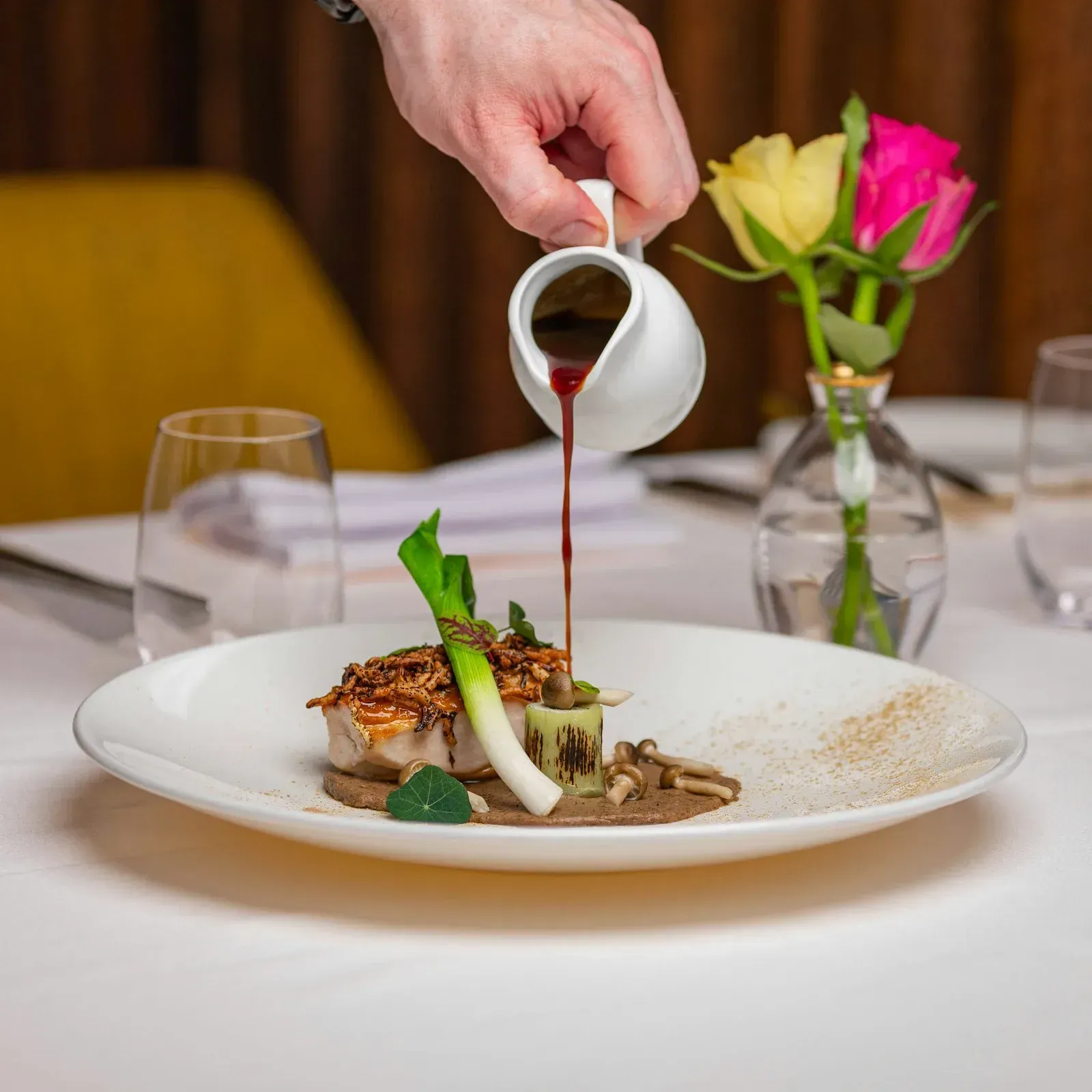 A chef pouring sauce over an artfully plated dish with edible flowers.